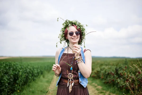 Young hippie woman with short red hair, wearing boho style clothes and flower wreath, standing on green currant field, posing for picture. Female portrait on natural background. Eco tourism concept.