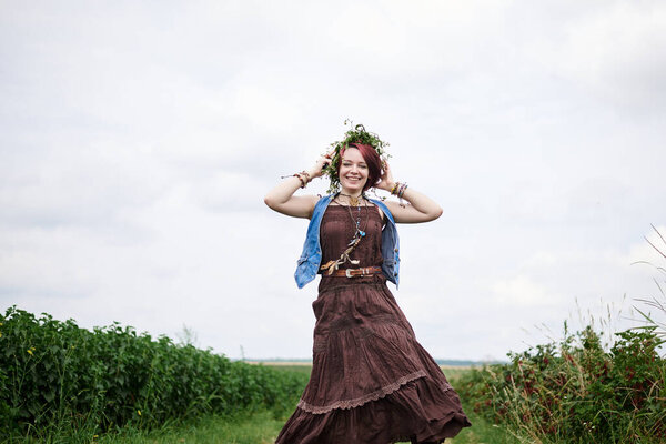 Young hippie woman with short red hair, wearing boho style clothes and flower wreath, running jumping on green field, smiling, laughing. Female portrait on natural background. Eco tourism concept.