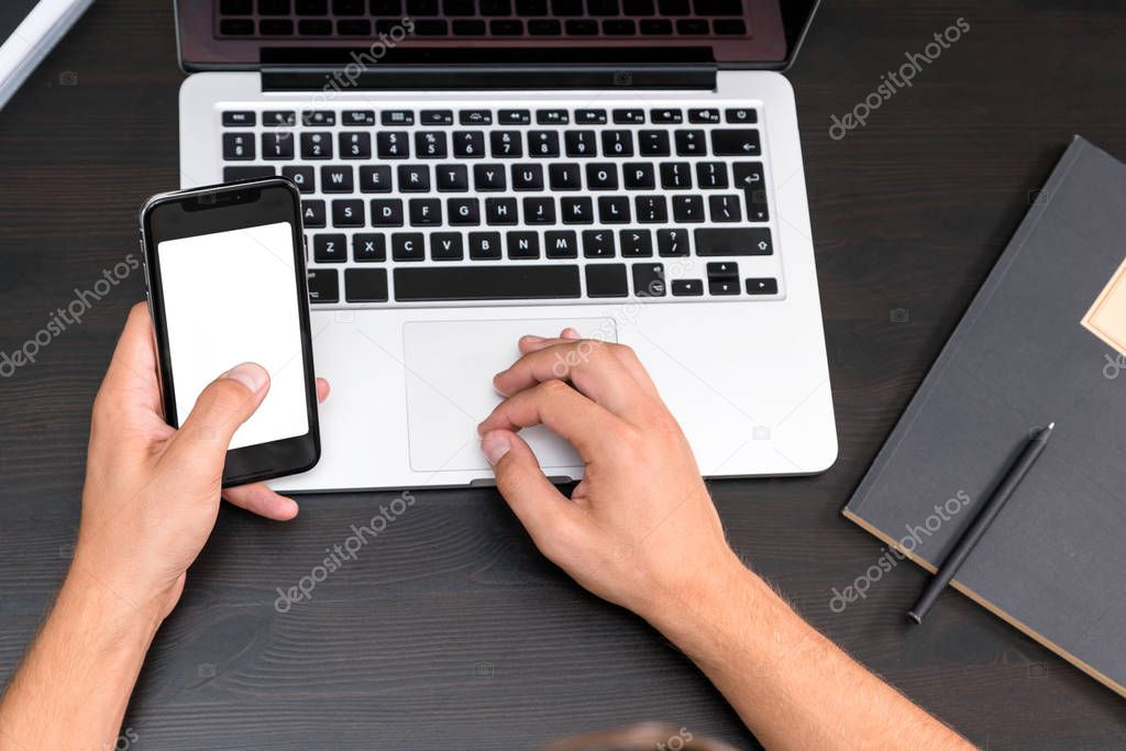 Mans hands typing on smart phone while working on laptop computer on vintage wooden table. Hands typing on a keyboard. Top view, business office workplace