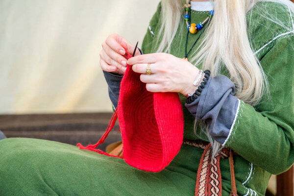 Close-up of woman hands knitting red wool hat, while wearing rural linen clothing. Freelance creative working and living concept