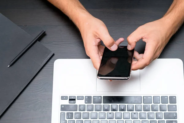 Mans hands typing on smart phone while working on laptop computer on vintage wooden table. Hands typing on a keyboard. Top view, business office workplace