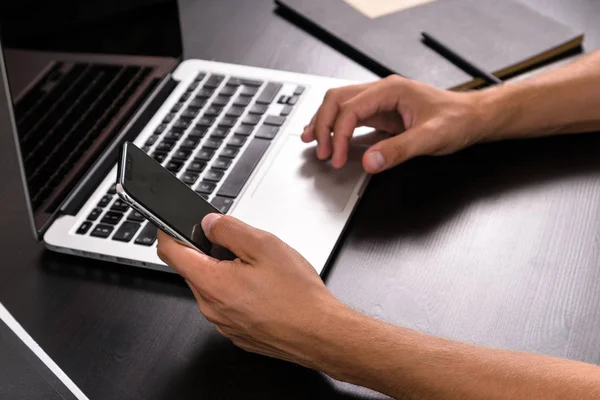 Mans hands typing on smart phone while working on laptop computer on vintage wooden table. Hands typing on a keyboard. Top view, business office workplace