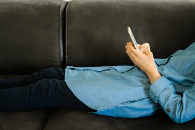 Elinde boş bir ekran ile bir akıllı telefon tutarken bir kanepeüzerinde yatan mavi elbiseli genç kız. Genç kız sosyal medyada kaydırma kullanarak, gençlik bağımlılığı