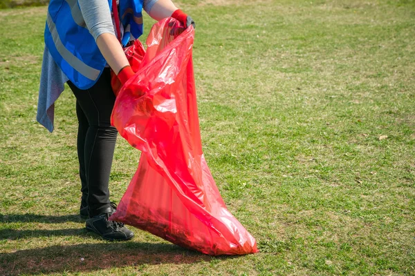 Woman volunteer wearing picking up trash and plastic waste in public park. Young girl wearing gloves and putting litter into red plastic bag outdoors