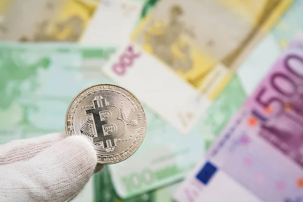 Hombre con clavo de olor blanco sosteniendo Bitcoin entre los dedos con billetes de banco euro en el fondo. Moneda digital, mercado de cadena de bloques — Foto de Stock