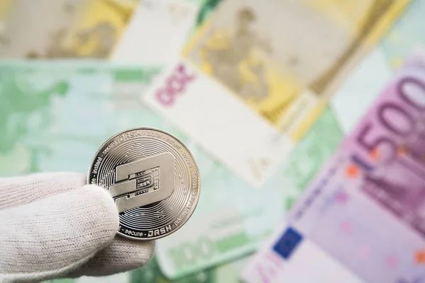 Hombre con clavo blanco sosteniendo moneda Dash entre los dedos con billetes de banco en euros en el fondo. Moneda digital, mercado de cadena de bloques — Foto de Stock