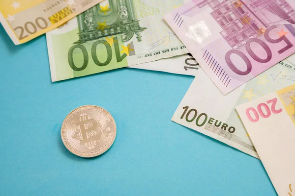 Bitcoin de plata junto a los billetes de banco de euros sobre fondo azul. Moneda digital, mercado de cadena de bloques. Billetes de euros junto a la moneda criptográfica — Foto de Stock