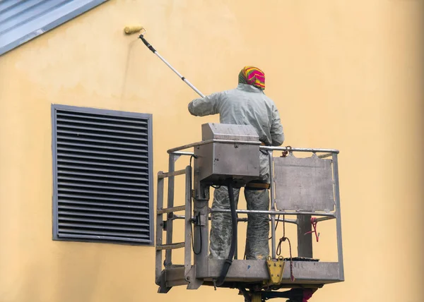 Man on a lifting platform painting the building wall with a roller exterior outdoors. Worker on a ladder manually painting yellow wall on construction site — Stock Photo, Image