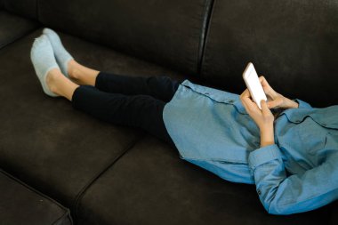Elinde boş bir ekran ile bir akıllı telefon tutarken bir kanepeüzerinde yatan mavi elbiseli genç kız. Genç kız sosyal medyada kaydırma kullanarak, gençlik bağımlılığı