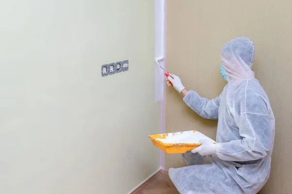 Man in protective work clothes painting the walls white colour with a paint roller. Home renovation project. Apartment renewal concept.