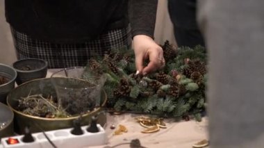 Noel çelengi dokuma atölyesi. Kadın, masa üzerinde ladin dalları, koniler ve çeşitli organik süslemelerden oluşan bayram çelenklerini elleriyle süslüyor.