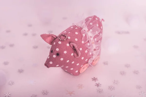 Handmade pink pig with white polka dots and skirt stands on white pink background snowflakes