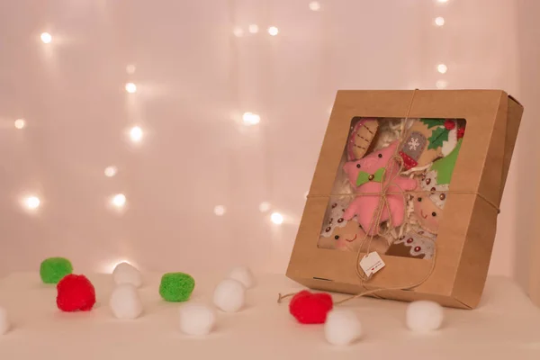 a box with a set of felt toys made of pigs, dwarfs, stars and handmade cookies stands against the background of Christmas lights and soft balls