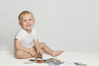 A toddler boy sits on a rug, laughs and plays with pills and medicines on a white background. clipart