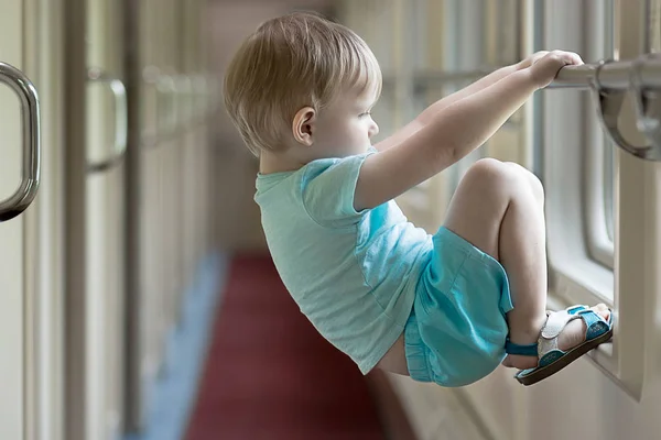 Znuděný a smutný blonďák toddler v modrých šatech visí na příčné liště okna vlaku. — Stock fotografie