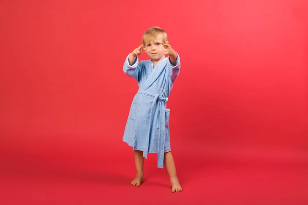 Blonďatý Chlapec Modrém Županu Červeném Jasném Pozadí Ukazuje Znak Ruky — Stock fotografie