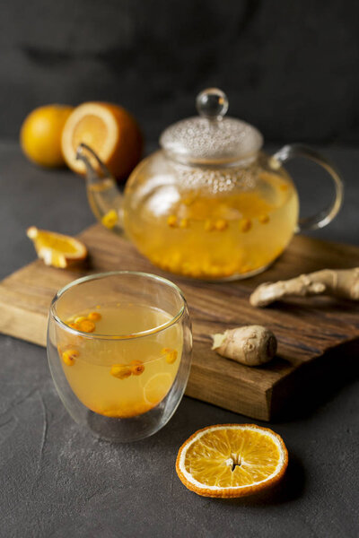 Hot sea buck thorn tea in glass teapot with glass cup, orange and ginger near, at wooden board on black background