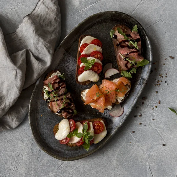 Sığır eti ve roka, somon ve kimyon, mozzarella ve domates kahverengi ekmek gri plaka üzerinde sandviç ile kızartma