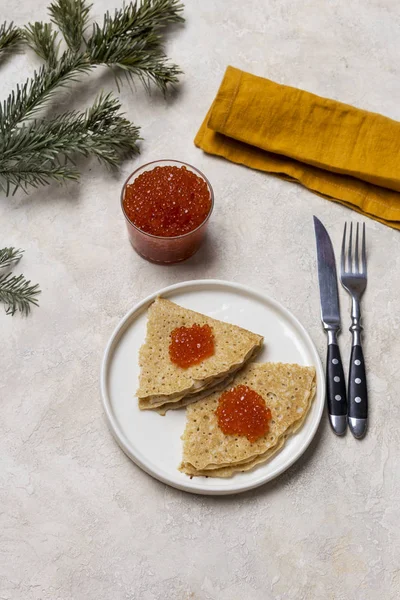Thin Pancakes with red caviar at white plate with jar of red caviar, fork, knife, napkin and fir near at light background