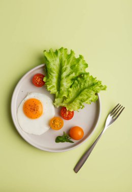Yeşil arka planda sağlıklı bir kahvaltı. Tuzlu ve biberli sahanda yumurta, kiraz domatesleri ve açık yeşil arka plan ve çatallı yeşil salata. Üst manzara, düz yatak, kıtasal kahvaltı konsepti.