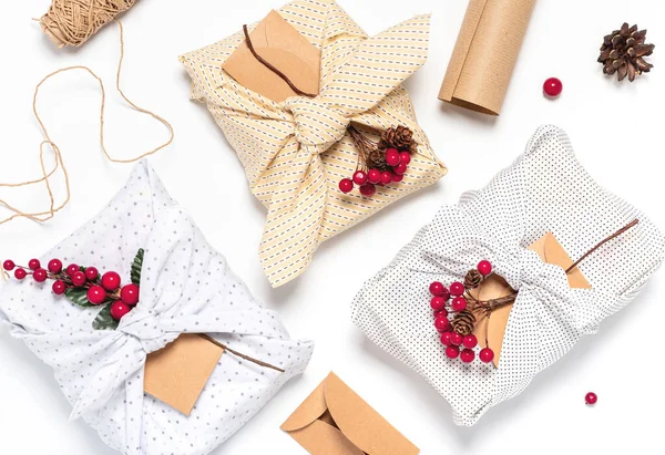 Christmas gifts wrapped in light textile and decorated Christmas branches with berries, flat lay. Christmas gifts in eco-friendly packaging on a white background, top view