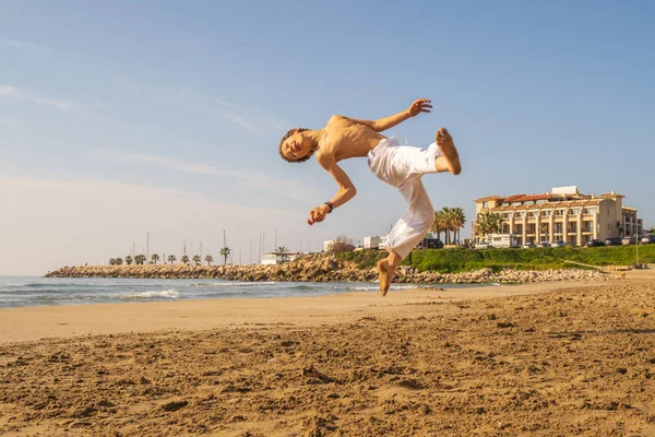 Boy in white pants practicing capoeira (brazilian martial art that combines elements of dance, acrobatics and music) on the beach