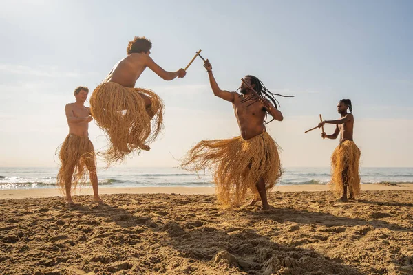 Men in maculele skirt practicing capoeira (brazilian martial art that combines elements of dance, acrobatics and music) on the beach