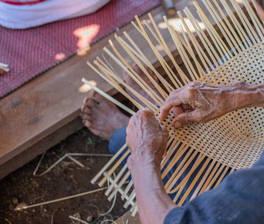 Becerikli, yaşlı, engelli bir adam yerde bambu hasır işi yapıyor. Kendine güvenme pratiğiyle basit bir yaşam tarzı gösteriyor..