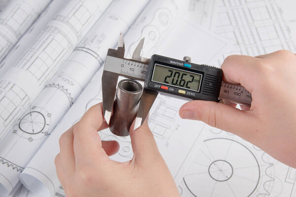 Engineer work. Measurement of the metal part with a caliper on the background of engineering drawings