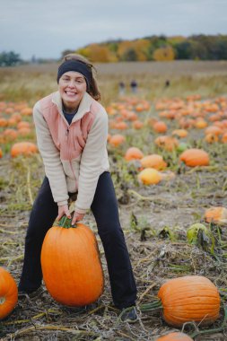 Adult woman (30s) attempts to pick up a giant pumpkin from a pumpkin patch. Smiling and laughing and having fun clipart