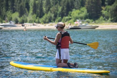 Active fit woman (30s) kneels on a stand up paddleboard on Lake Tahoe in the summer. clipart