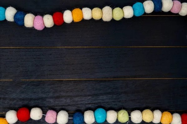 Pretty colorful rainbow pom pom fabric garland over black wood background. Useful for birthday projects or non traditional Chistmas backgrounds