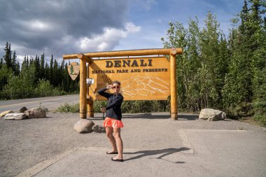 1 Ağustos 2018 - Denali Milli Parkı Alaska: bir selfie sopa ile onu smartphone Ördeksurat ifade için sosyal medya yapım park giriş işareti bir fotoğraf çekmek için bir turist kadın kullanır