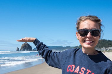 Woman wearing sunglasses pretends to touch a Haystack rock in Cannon Beach, Oregon on the beach on a sunny day. Forced perspective view clipart