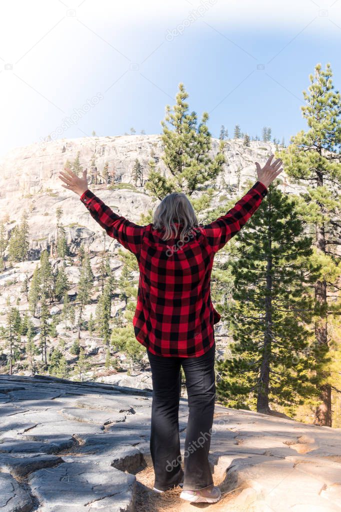 A mature blonde woman wearing a buffalo plaid flannel shirt (red and black) raises arms up in the Eastern Sierra Nevada mountains in California, on top of Devil's Postpile National Monument