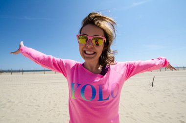 Young woman stands on the beach, wearing a neon pink YOLO shirt with sunglasses, as her hair blows in the wind clipart