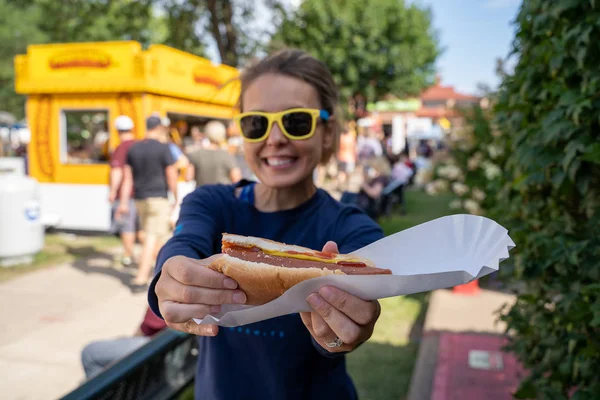 Smiling woman at an outdoor fair holds up a half-eaten footlong — Stock Photo, Image