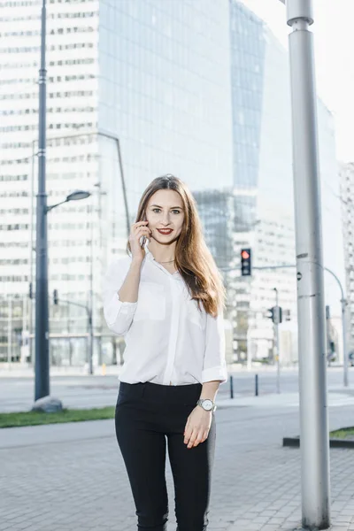 A business woman speaks through the phone. A good girl is sitting on the bench. A young woman in the city center.