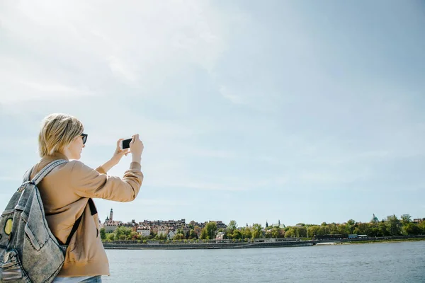 The girl picks up the city over the river on the phone. Young woman with a mobile phone near the river. Panorama of the city. Mobile photography. stylish woman with shoulder bag and sunglasses on the beach near Wisla. The lady travels to Warsaw. Euro