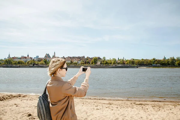 The girl picks up the city over the river on the phone. Young woman with a mobile phone near the river. Panorama of the city. Mobile photography. stylish woman with shoulder bag and sunglasses on the beach near Wisla. The lady travels to Warsaw. Euro