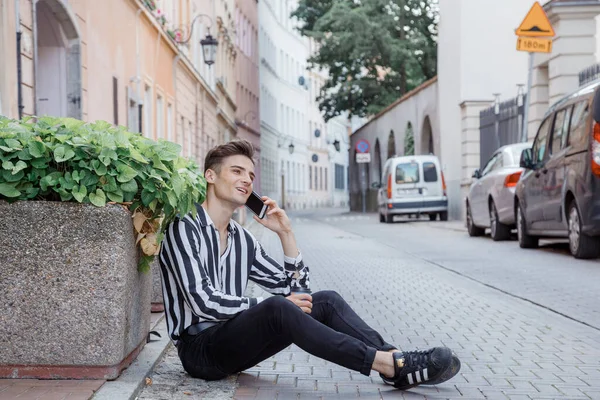 A young guy in a striped shirt sits on the road near a flower bed with pink flowers and talks on the phone. Stylish young man sitting on an old city street. Traveling through the cities of Europe.