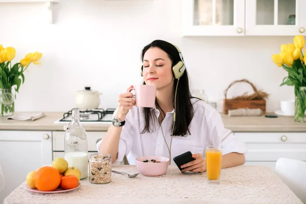 Young beautiful woman using enjoying music and coffeewhile healthy breakfast in light modern kitchen.