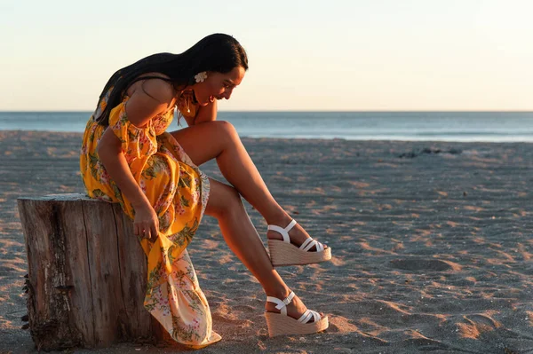 Sarı elbiseli zarif bir beyaz kadın manken ve kumsalda bir kütükte otururken gülümseyen beyaz takoz.