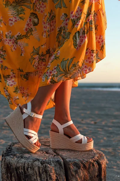 Sarı elbiseli, çiçek desenli ve plajda zarif beyaz takozlu bir kadının bacakları.