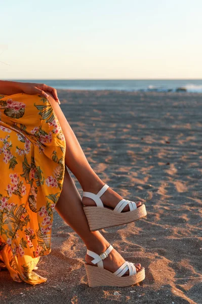 Sarı elbiseli, çiçek desenli ve plajda zarif beyaz takozlu bir kadının bacakları.