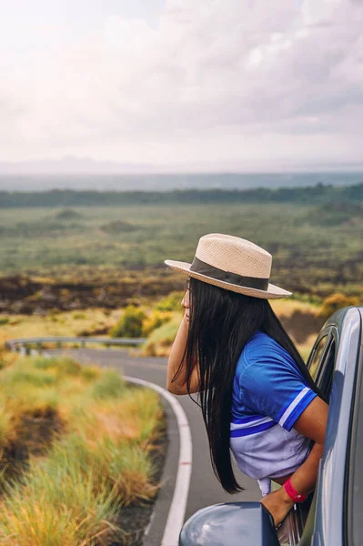 Arabanın camından sarkan şapkalı genç bir kadın. Arka planda güzel bir manzara var.