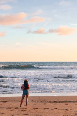 Gün batımında denizin dalgalarının önünde duran genç bir kadının arka görüntüsü.
