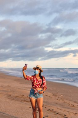 Çiçek desenli, şortlu ve şapkalı güzel kırmızı bir gömlek giyen genç bir bayan, deniz kıyısında yüz maskesi takarak covid-19 'u önlemek için akıllı telefon kullanıyor. Coronavirus salgını sırasında tatil. Yeni normal kavram.