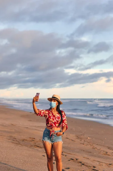Çiçek desenli, şortlu ve şapkalı güzel kırmızı bir gömlek giyen genç bir bayan, deniz kıyısında yüz maskesi takarak covid-19 'u önlemek için akıllı telefon kullanıyor. Coronavirus salgını sırasında tatil. Yeni normal kavram.