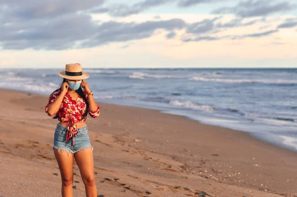 Çiçek desenli, şortlu ve şapkalı güzel kırmızı bir gömlek giyen genç bir kadın, deniz kıyısında dikiliyor ve kovid-19 'u önlemek için maske takıyor. Coronavirus salgını sırasında tatil. Yeni normal kavram.